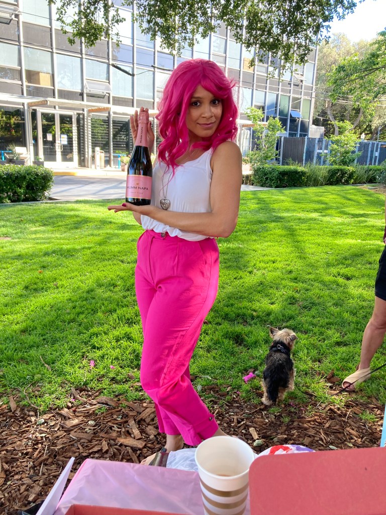 Addie Woolridge wearing a bright pink wig and bright pink pants. She is holding a bottle of Mumm Napa