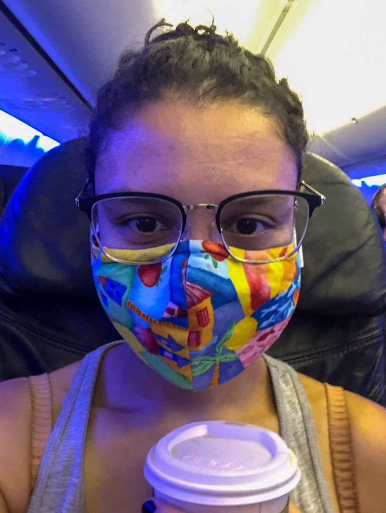 A very close up photo of Addie. Her hair is pulled back and she is wearing glasses and a brightly colored mask and holding a cup of coffee