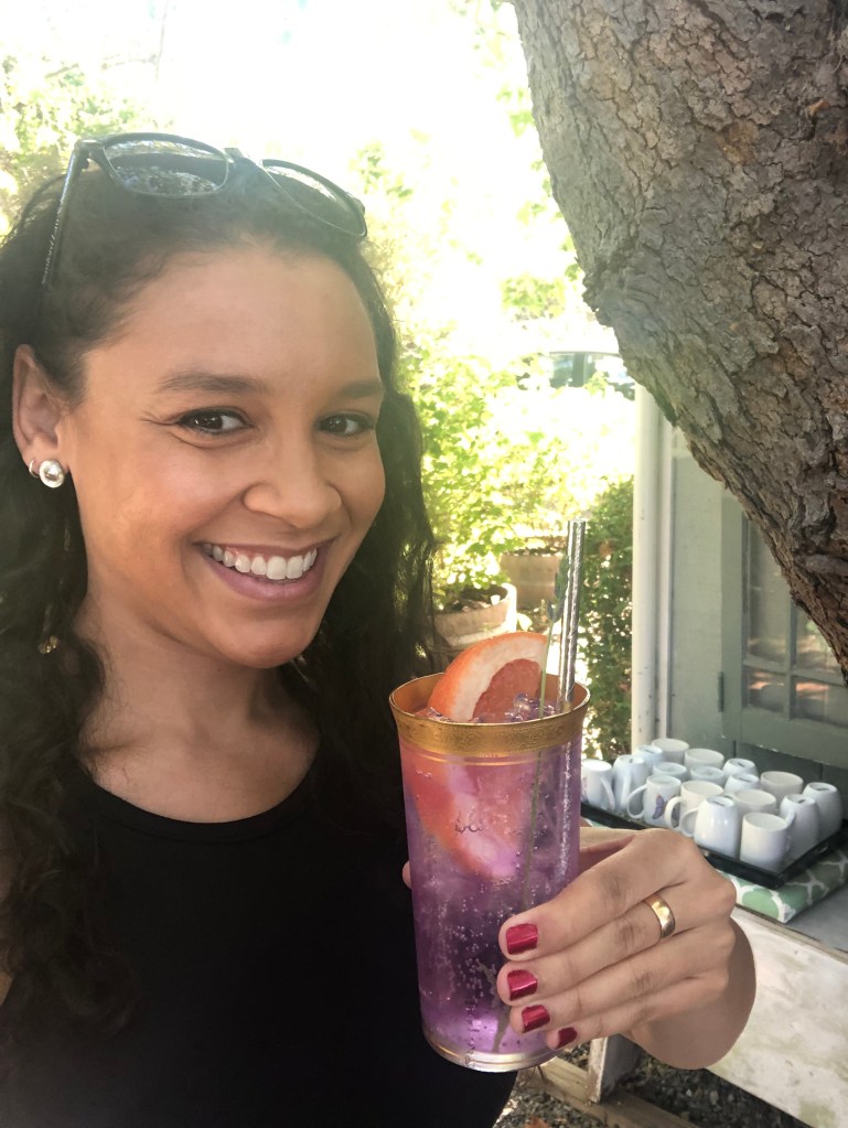 Addie Woolridge holding a cocktail that is purple with an orange slice and silver straw. She is wearing black with sunglasses on the told of her head and her nail polish is red.