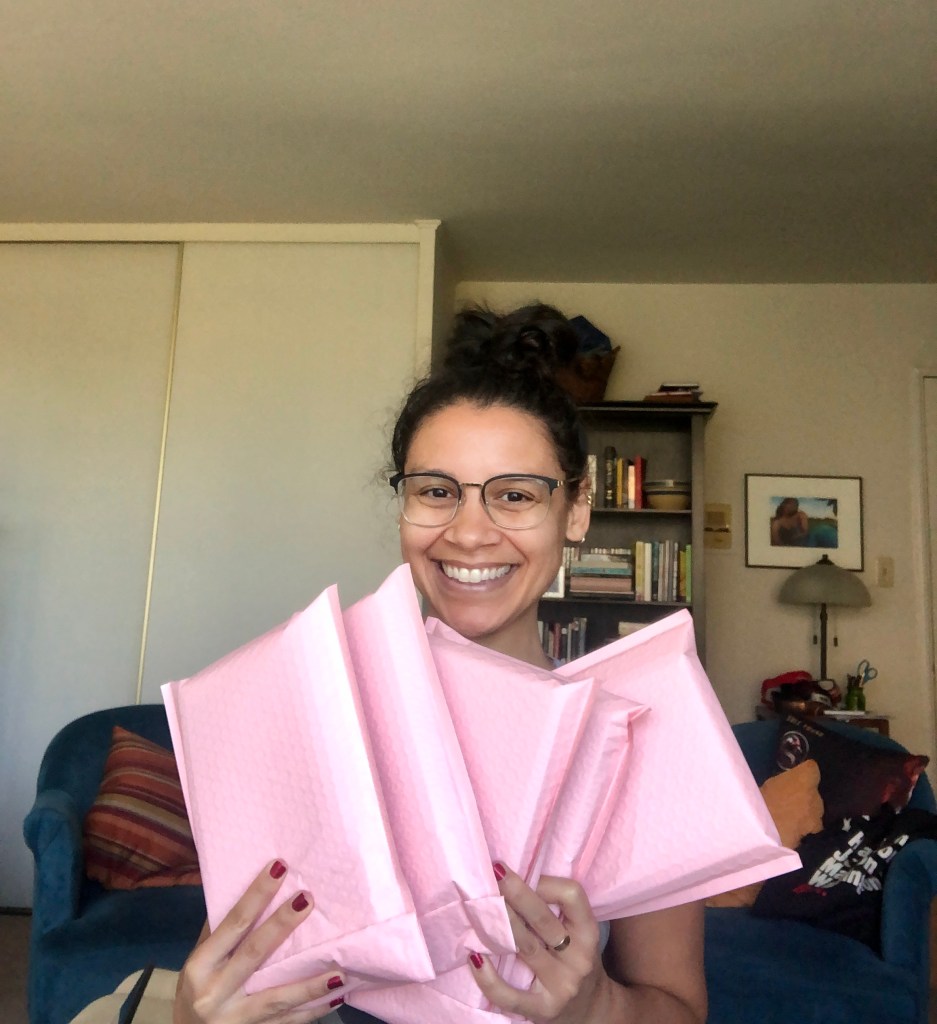 Addie holding a bunch of pink packages containing books. She is wearing glasses and her hair is in a messy bun. Her nail polish is red.