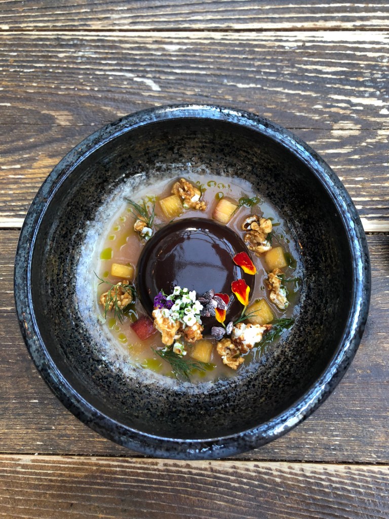 A chocolate desert in a blue bowl, covered in flowers and pieces of citrus fruit.