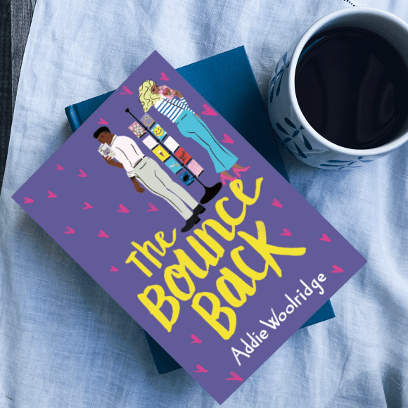 The book The Bounce Back sits on a pile of books on top of a white table cloth with a mug of coffee next to it.