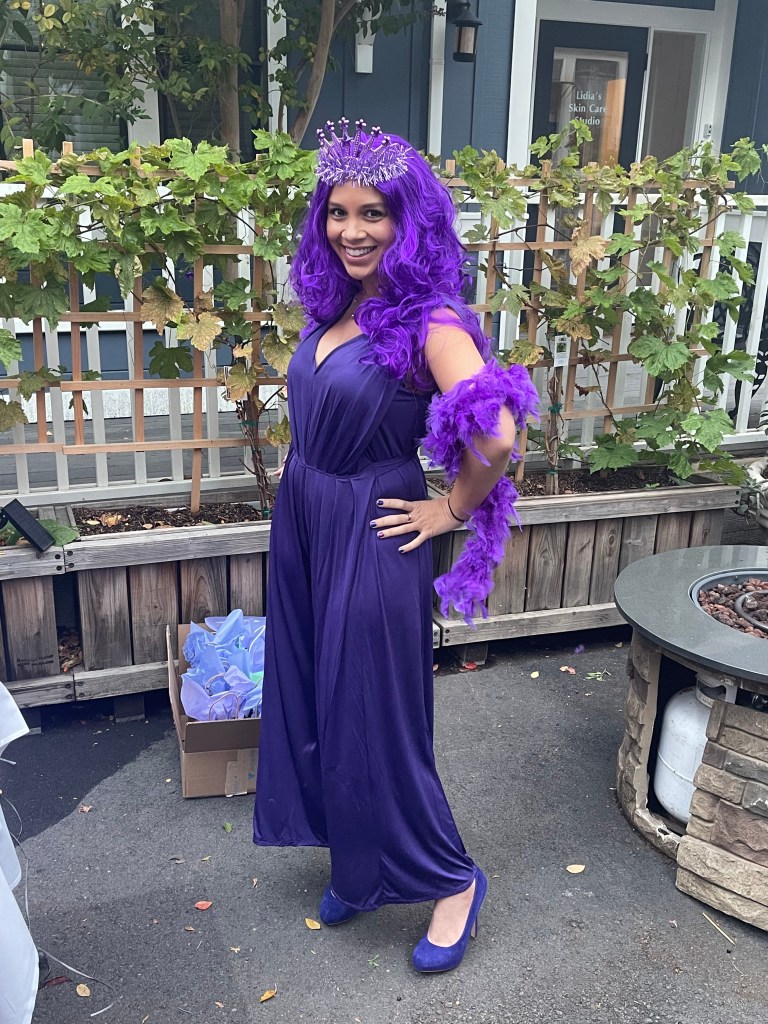 Addie Woolridge is standing with her hands on her hips and smiling. She is wearing a purple jumpsuit, boa and heels and a purple wig with a crown.