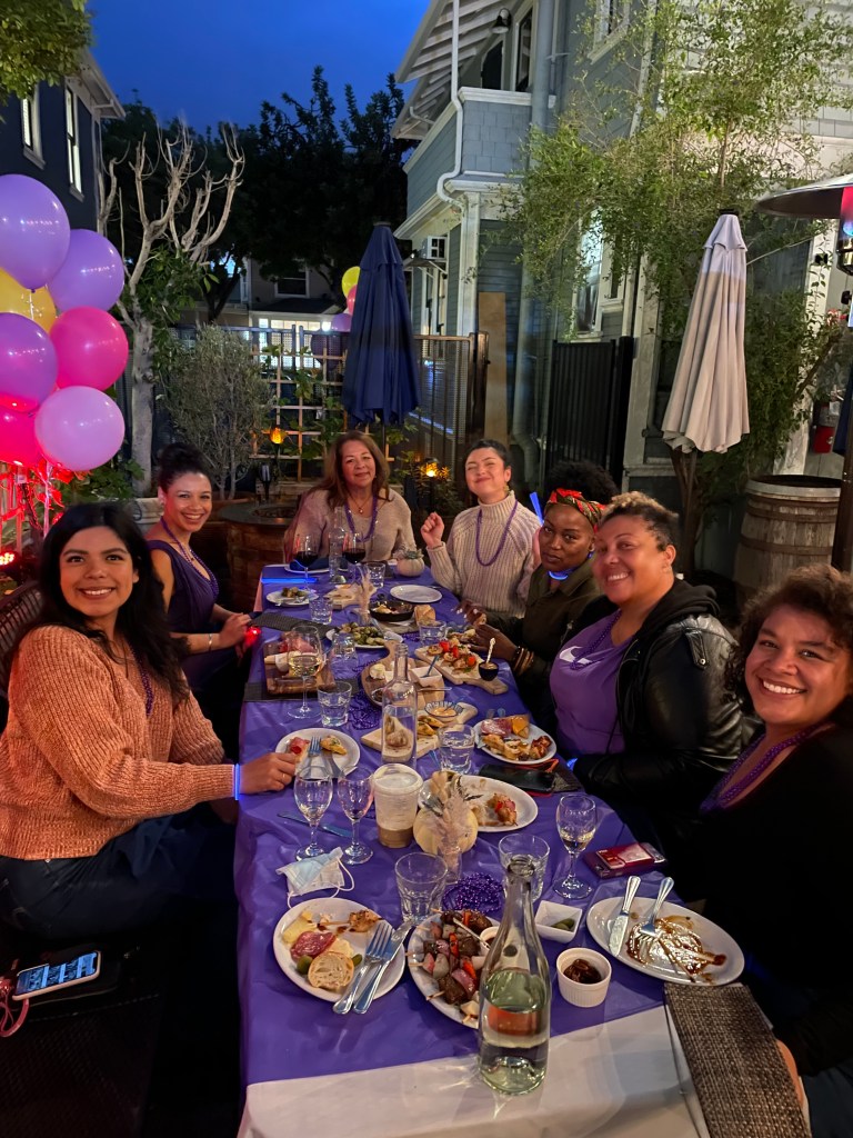 A group of seven friends is seated around a long table enjoying a meal and smiling. The table is white with purple linens and there are purple balloons in the background.