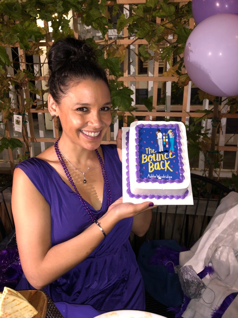Addie Woolridge is wearing a purple jumpsuit. Her hair is up in a bun and she is smiling while holding up a rectangular cake with white and purple frosting and a picture of The Bounce Back on it. 