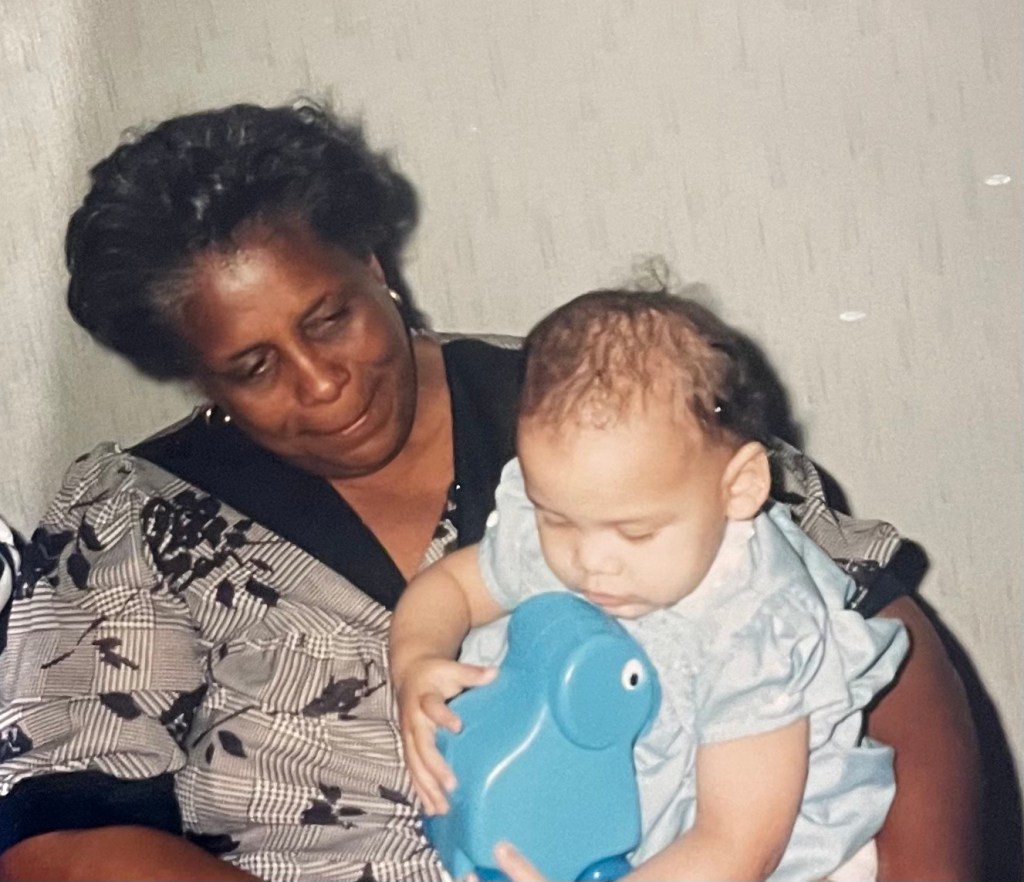 Me and my Grandmama on my first birthday. She is Black and has short black hair that is streaked with grey. She is smiling and I am holding a blue plastic toy